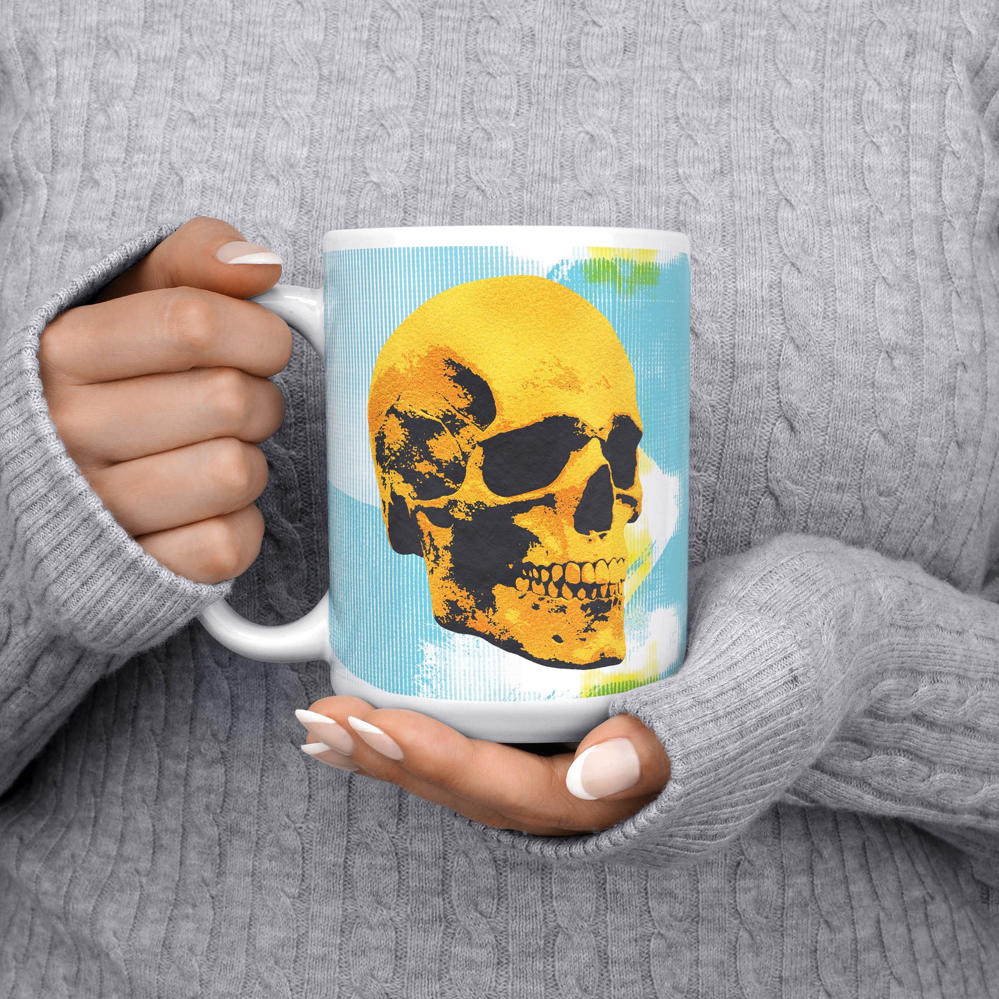 Close-up of a woman in a gray pullover holding the 11oz Golden Skulls High Five coffee mug, side view with the handle on the right, showcasing the detailed pop art design of two golden skulls high-fiving on a light blue background.