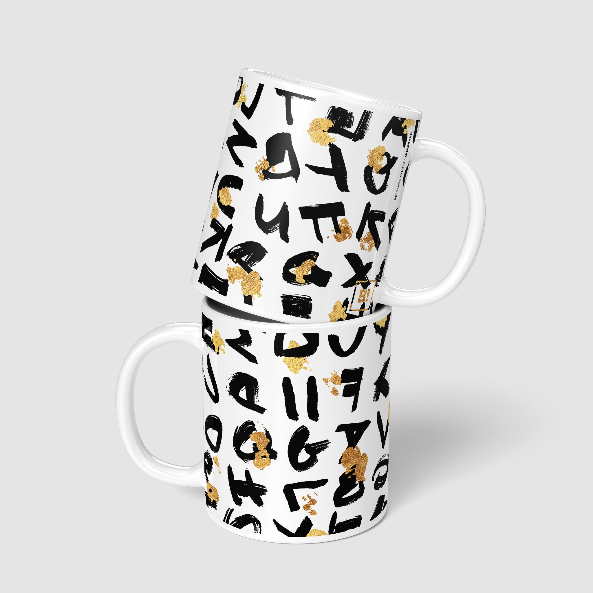 Two 11oz coffee mugs stacked, one on top of the other, revealing both sides of the design with black random lettering and golden splatter, symbolizing a blend of randomness and elegance.