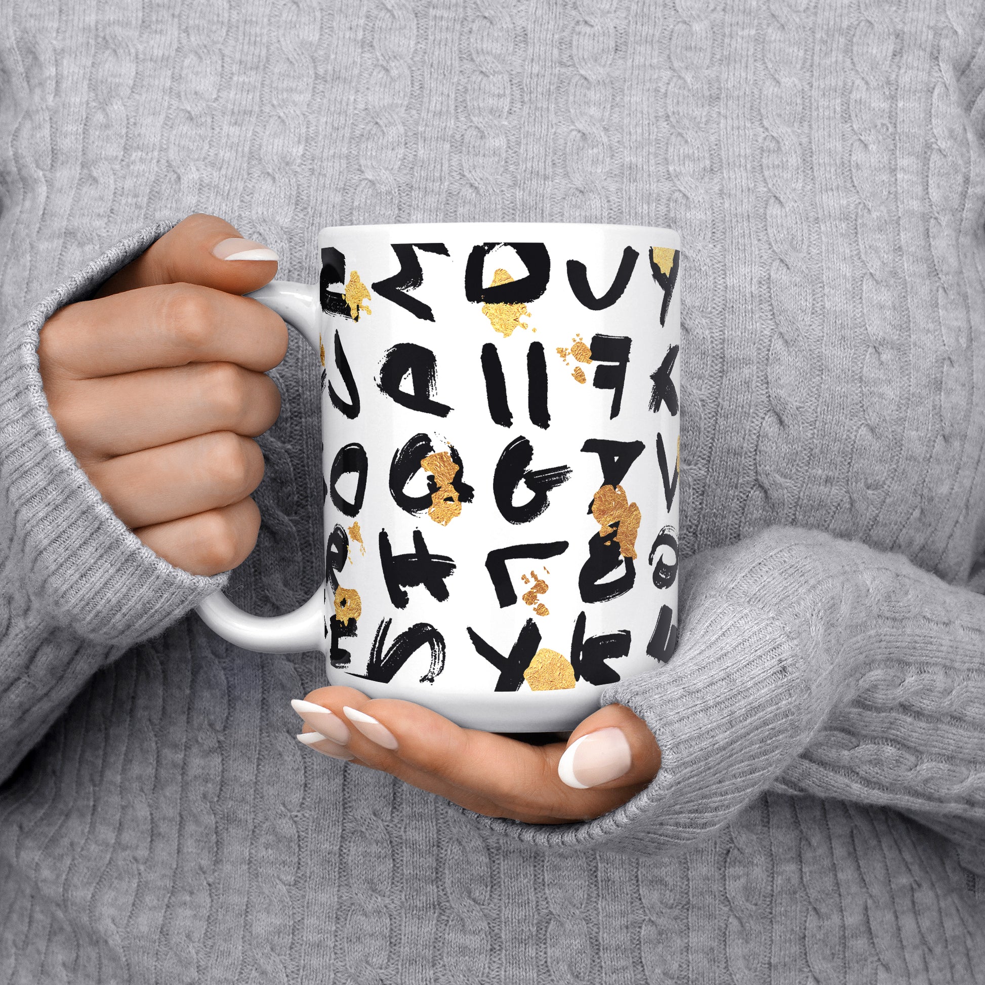 Close-up of a woman in a gray pullover holding the 11oz coffee mug, side view with handle on the right, showcasing the artistic black random lettering and golden splatter design.